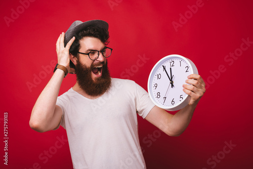 Young bearded hipster holding a white clock and watching it shocked, screaming and stressed on red background.