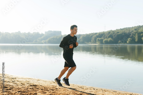 Young man is running on the shore of lake in the park in the morning wearing black sportswear.