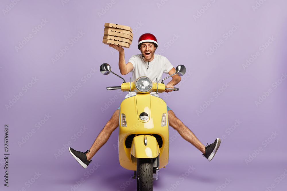 Busy deliveryman being in hurry, carries cardboard boxes with pizza, delivers to customers, poses on yellow scooter, wears helmet, white t shirt and sportshoes, spreads legs, has happy expression - obrazy, fototapety, plakaty 