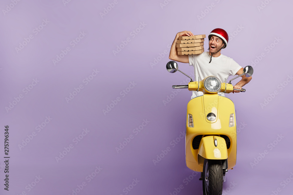 Happy male courier rides motorbike, keeps pile of boxes with delicious pizza, delivers to people and does his duties during work, isolated on purple wall, free space on left. Fast delivery concept - obrazy, fototapety, plakaty 