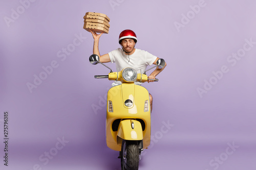 Nervous male courier distributes pizza in cardboard containers on motorbike, tries to reach destination quickly and be punctual, being professional well skilled driver, stuck in traffic jam on his way