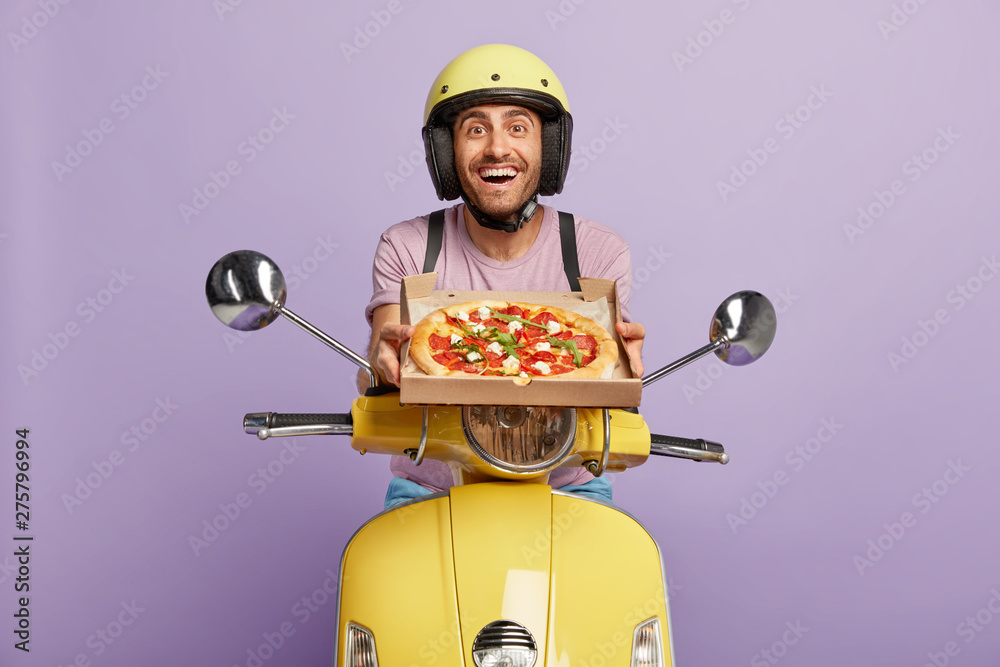 Friendly looking punctual pizzaman has good time management skills, arrives always in time, shows delicious pizza for customer, hopes for getting tip, wears yellow helmet, rides on fast motorcycle - obrazy, fototapety, plakaty 