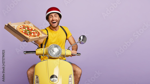 Bilde på lerret Cheerful male restaurant worker holds opened box of delicious tomato pizza, offers to taste, has broad friendly smile, rides motorbike, delivers food free, isolated on purple wall