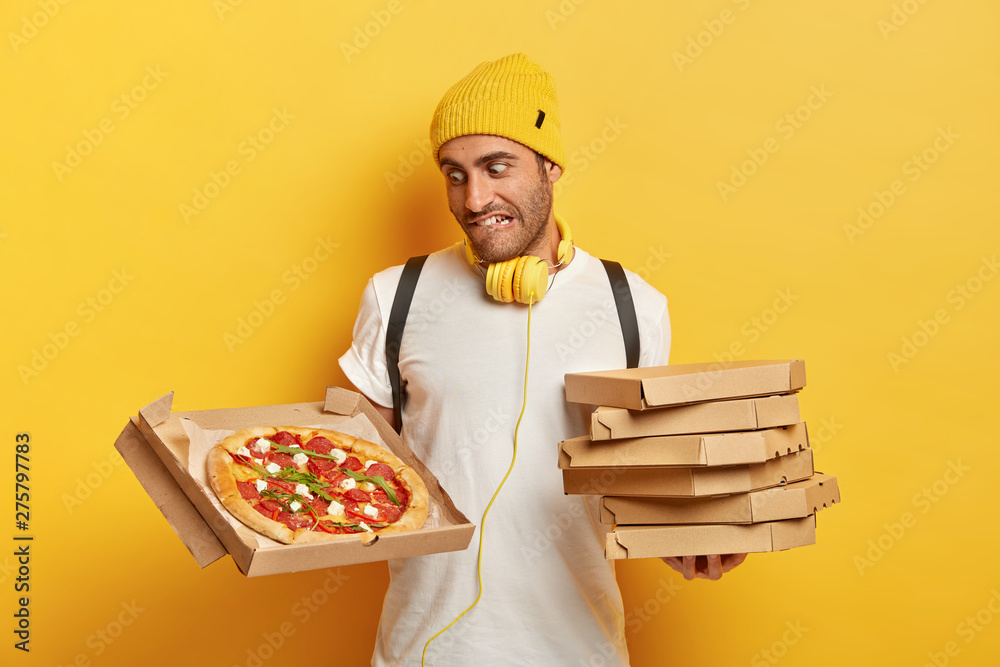 Handsome salesperson or courier represents delicious pizza, bites lips as looks at tasty junk food, holds stack of cardboard boxes, wears casual clothes isolated on yellow background. Delivery service Handsome salesperson or courier represents delicious pizza, bites lips as looks at tasty junk food, holds stack of cardboard boxes, wears casual clothes isolated on yellow background. Delivery service