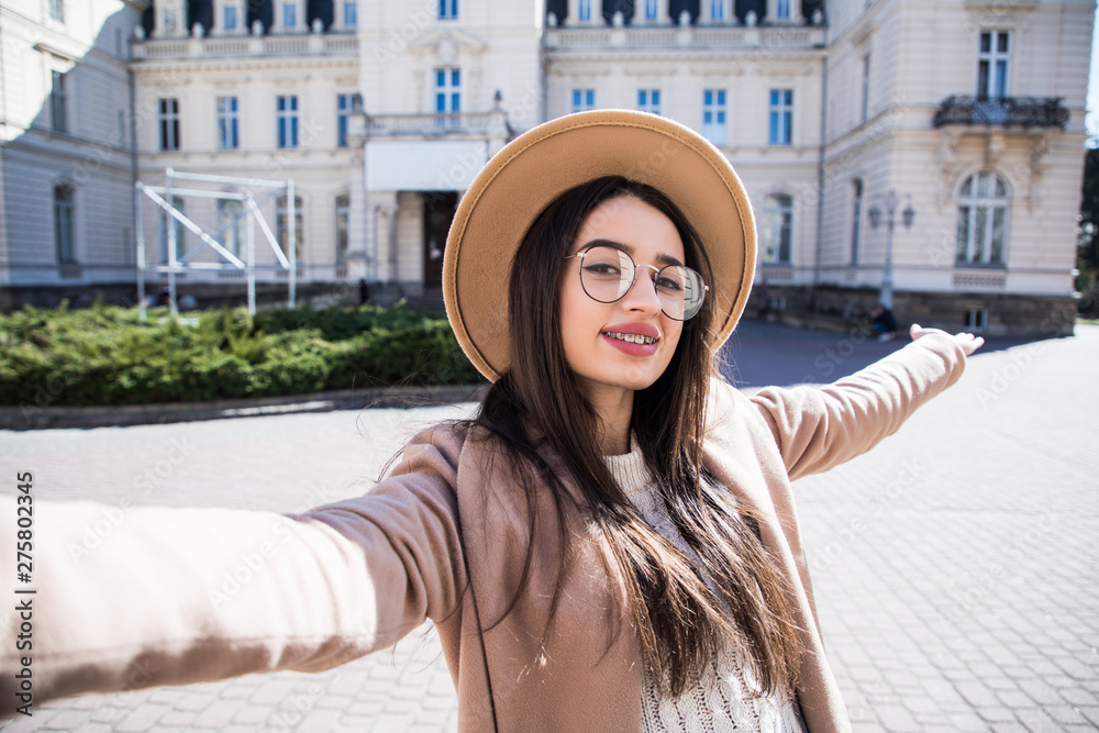 Young girl wear in coat and hat take selfie from hands with phone on summer city street. Urban life concept.