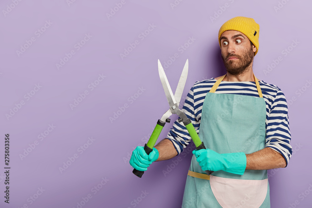 Photo of stupefied man gazes with shocked expression at pruning shears, involved in grooming, holds gardening tool, wears uniform, rubber gloves, isolated on purple wall. Agriculture concept Photo of stupefied man gazes with shocked expression at pruning shears, involved in grooming, holds gardening tool, wears uniform, rubber gloves, isolated on purple wall. Agriculture concept