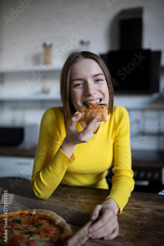 Funny brunette girl in yellow sweater eating pizza at kitchen