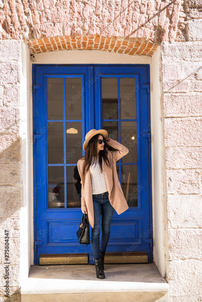 Young pretty woman woman wear in coat and hat with smile stands at the blue door on the street