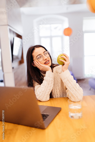 Portrait of a beautiful young student girl with apple sitting indoors using laptop computer.