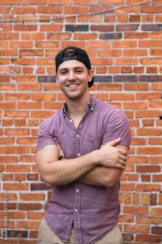 Handsome Young Caucasian Man with Backwards Hat Smiling for Portraits in Front of Textured Brick Wall Outside