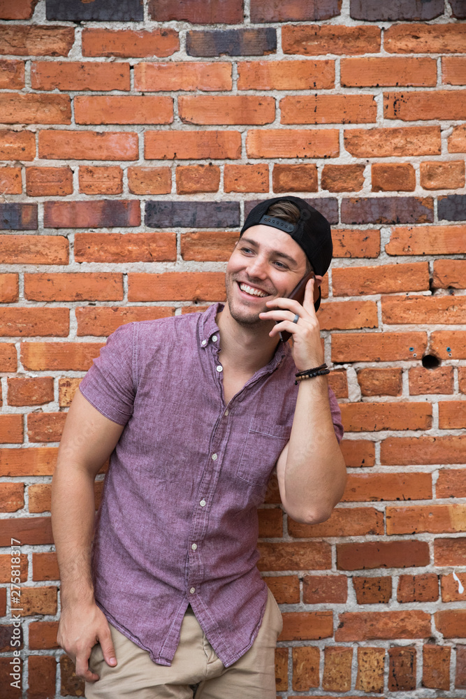 Handsome Young Man on Cellphone Texting and Call Conversation with Backwards Hat Smiling for Portraits in Front of Textured Brick Wall Outside Handsome Young Man on Cellphone Texting and Call Conversation with Backwards Hat Smiling for Portraits in Front of Textured Brick Wall Outside