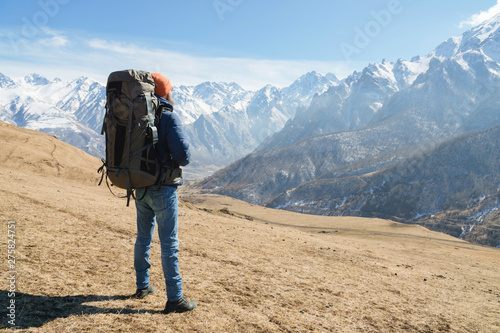 A bearded man in a hat and a backpack is standing outdoors against the snowy mountain peaks on a sunny day. Back view