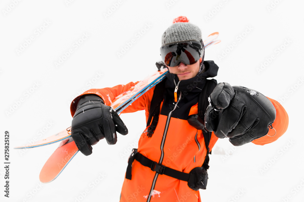 Professional skier athlete in a knitted hat and orange black suit with a black ski mask with skis on his shoulder points to the camera during a snowstorm on a light background in the snow. Professional skier athlete in a knitted hat and orange black suit with a black ski mask with skis on his shoulder points to the camera during a snowstorm on a light background in the snow.