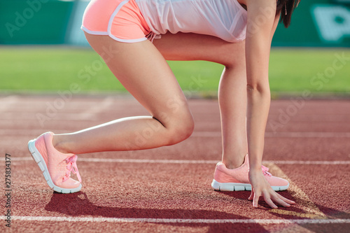 Young sporty woman in pink shorts and tank tops are ready to run and stands on start on stadium track. Close-up legs and arms