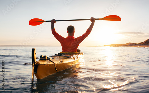 Rear view of active senior man holds kayak paddles high toward sun.