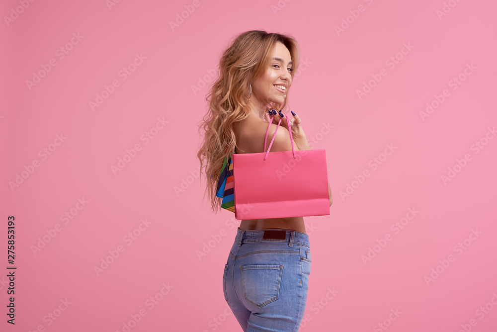 Portrait of an excited beautiful smiling girl wearing colorful clothes holding shopping bag isolated over pink background. Place to sign or text on the package - obrazy, fototapety, plakaty 