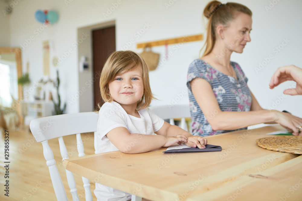 Portrait of cute adorable baby girl in white t-shirt sitting at wooden dining table wiht her mother, learning how to make origami paper plane, looking at camera with happy smile. Selective focus Portrait of cute adorable baby girl in white t-shirt sitting at wooden dining table wiht her mother, learning how to make origami paper plane, looking at camera with happy smile. Selective focus