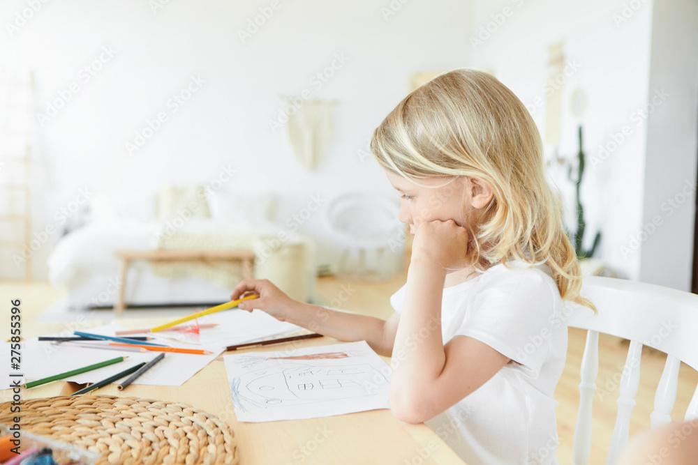 Horizontal image of creative talented little European child with loose blonde hair sitting at wooden desk in stylish bedroom interior with sheets of paper and colorful pencils, drawling and painting Horizontal image of creative talented little European child with loose blonde hair sitting at wooden desk in stylish bedroom interior with sheets of paper and colorful pencils, drawling and painting