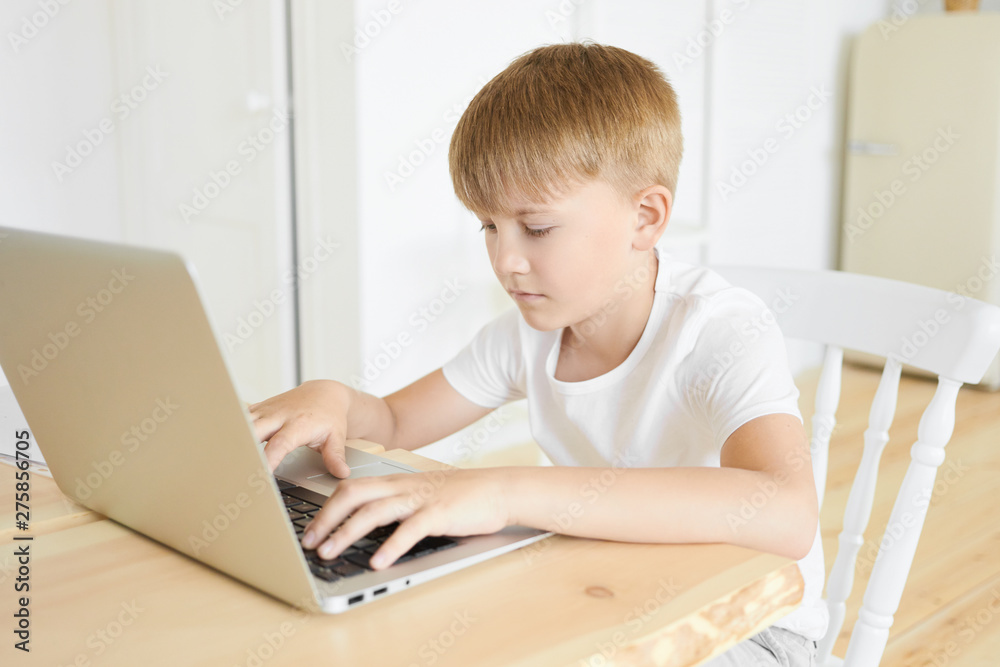 Portrait of handsome serious Caucasian boy of school age sitting at wooden table using laptop computer, keeping hands on keyboard. Education, leisure, people and modern electronic gadgets concept Portrait of handsome serious Caucasian boy of school age sitting at wooden table using laptop computer, keeping hands on keyboard. Education, leisure, people and modern electronic gadgets concept