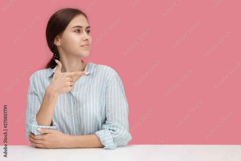 Horizontal shot of beautiful joyful young in stylish striped blue shirt working at office desk and making gesture, pointing index finger at blank pink wall with copyspace for your promotional content Horizontal shot of beautiful joyful young in stylish striped blue shirt working at office desk and making gesture, pointing index finger at blank pink wall with copyspace for your promotional content