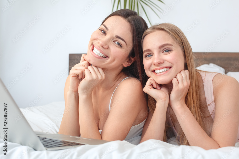 Friends together at home lying on bed with laptop posing to camera joyful Friends together at home lying on bed with laptop posing to camera joyful
