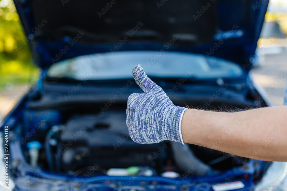 Hands in grey gloves of professional young mechanic man showing thumb up, okey as sign of success with car in open hood at the garage background. Auto repair service. Hands in grey gloves of professional young mechanic man showing thumb up, okey as sign of success with car in open hood at the garage background. Auto repair service.