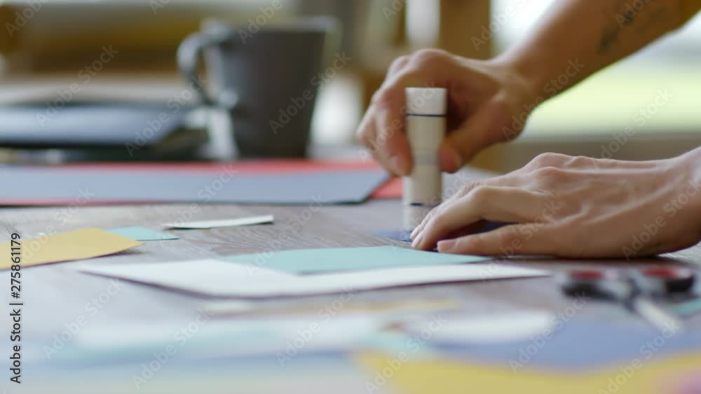 Close up hands of unrecognizable young woman using glue stick to attached colored paper pieces to collage background Close up hands of unrecognizable young woman using glue stick to attached colored paper pieces to collage background