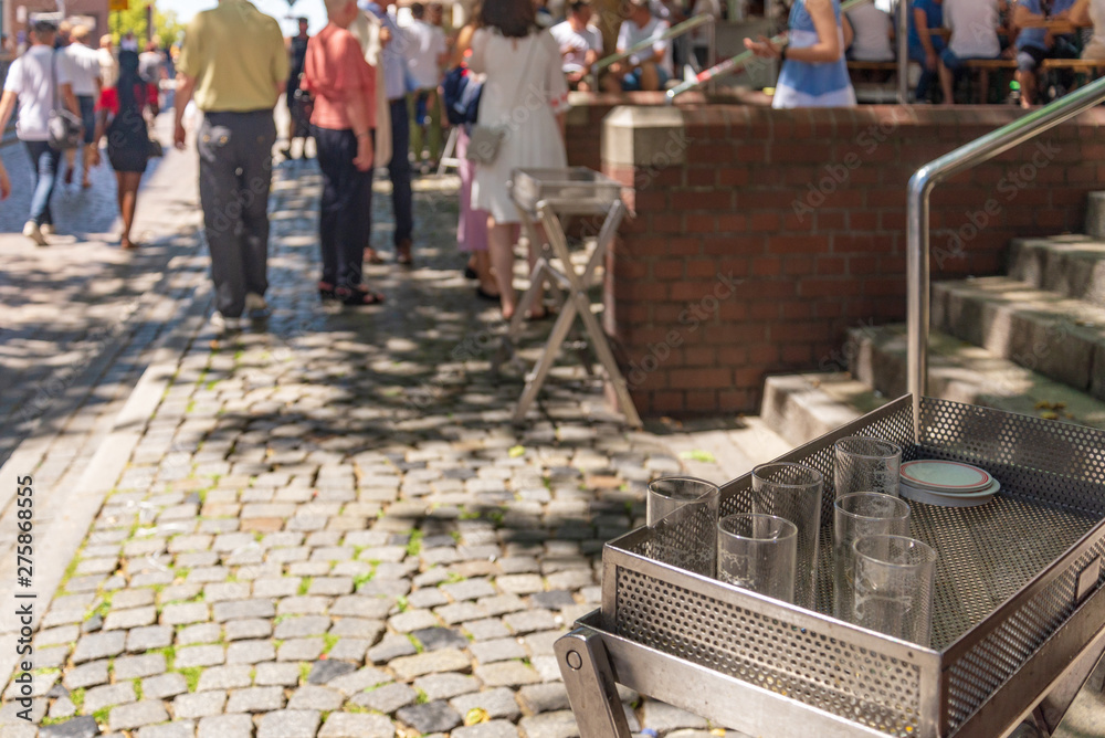 Outdoor sunny selected focus view at metal trays with fully glasses of beer stand on the walking street, and blur background of people stand, hang out and drink beer in old town Düsseldorf, Germany.  