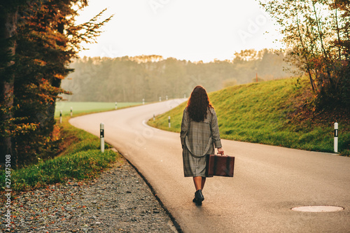 Beautiful woman walking away down the road, wearing long coat, holding old vintage suitcase, back view