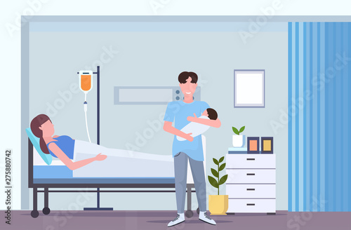 father holding newborn baby man visiting his wife lying in hospital bed with dropper happy family parenthood childbirth concept maternity ward interior flat full length horizontal
