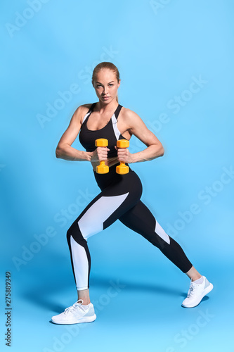strong slim girl doing lunges while holding dumbbell. isolated blue background. full length photo. lifestyle, hobby, free time
