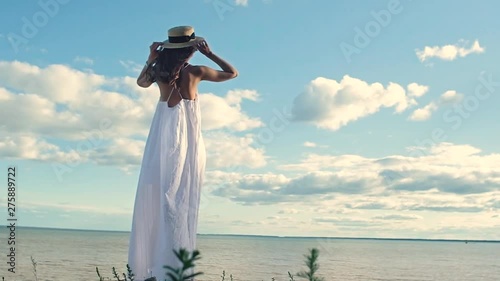 a beautiful slender girl in a white dress takes off her straw hat and spreads her hands to the sides, shakes her long hair and turns her face to the sun. The view from the back. Warm sunset on the sea