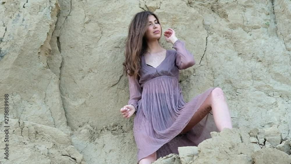 young attractive slender girl in a transparent brown dress and a black retro swimsuit, in dark shoes posing against the background of sandy gray cliff. Straightens his long hair and lifts his head up.