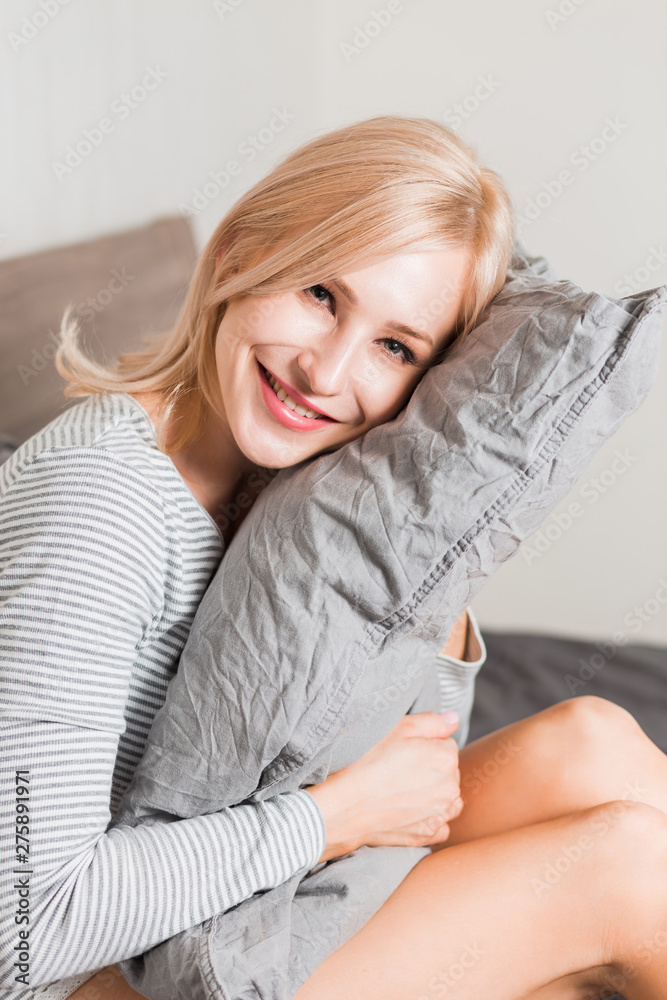 Positive woman with pillow sitting in bed and smiling to camera at home Positive woman with pillow sitting in bed and smiling to camera at home