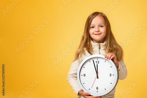 Cute little girl in a jacket holds a big clock on a yellow background. The concept of education, school, deadlines, time to study, autumn. Copy space