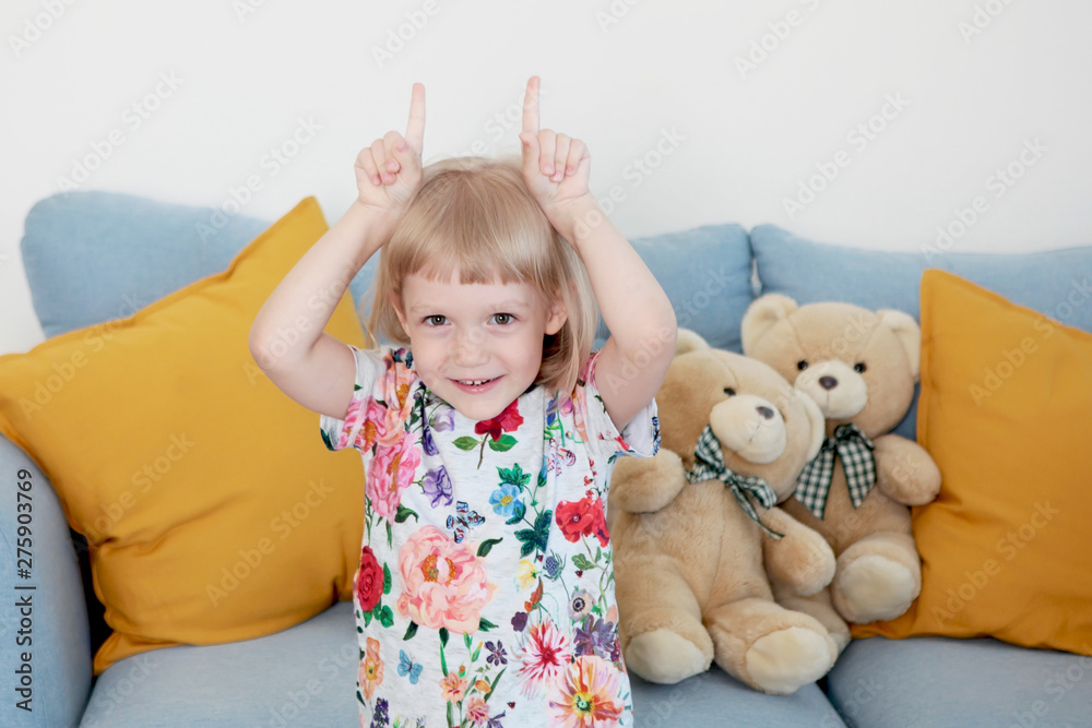 A little girl with blond hair is having fun, smiling, playing, showing horns on her head with her hands. Carefree childhood. The concept of a happy childhood. Photos in the interior. A little girl with blond hair is having fun, smiling, playing, showing horns on her head with her hands. Carefree childhood. The concept of a happy childhood. Photos in the interior.