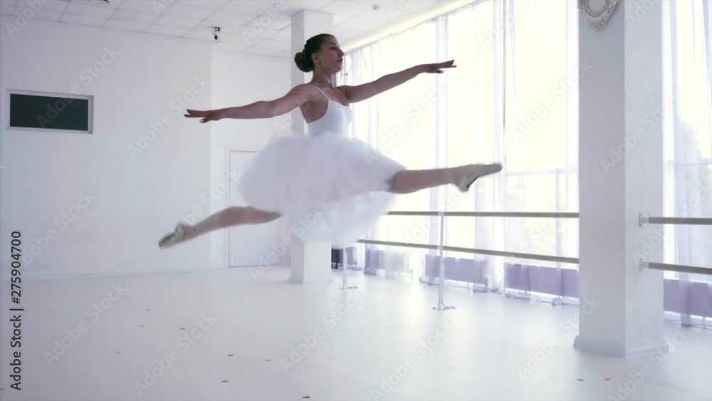 Ballet dancer is jumping up making grand temps leve passe. Classical dance in white tutu on pointe. She is performancing splits in the air. Young ballerina in light class with big windows.