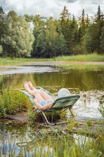 Middle aged woman in bikini, resting and sun bathing on sun chair by natural beautiful pond lake outdoors in summer day.