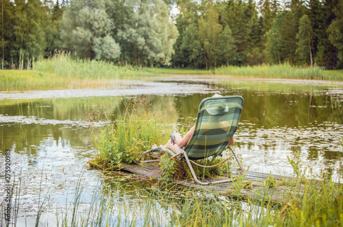 Middle aged woman in bikini, resting and sun bathing on sun chair by natural beautiful pond lake outdoors in summer day.