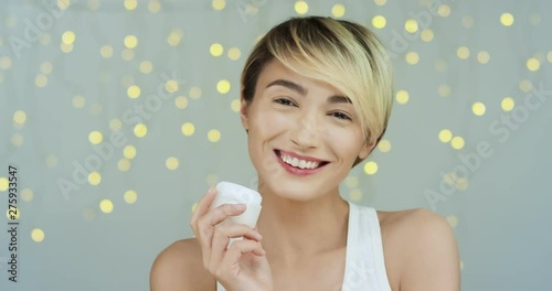 Portrait shot of the young Caucasian beautiful woman in the white shirt looking and smiling to the camera while holding a cream on hand and demonstating it to camera. photo