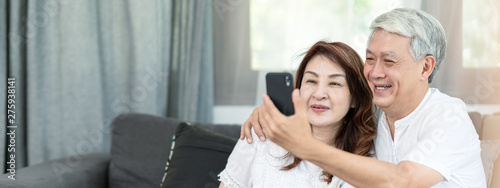Candid of attractive old asian couple husband and wife us technology for video call via smartphone 5G internet connection sitting on coach at home. Senior parent watching or looking at mobile screen.