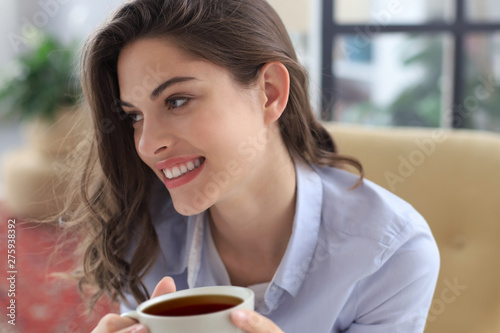 Smiling young female sitting in the armchair in the living room, holding a cup of coffee.