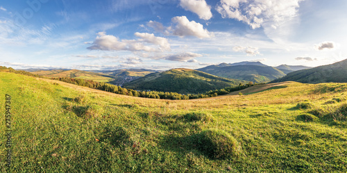 panorama of a countryside in mountains. beautiful early autumn landscape in evening light. grassy meadow on the hill. fluffy clouds above the distant ridge. village down in the valley