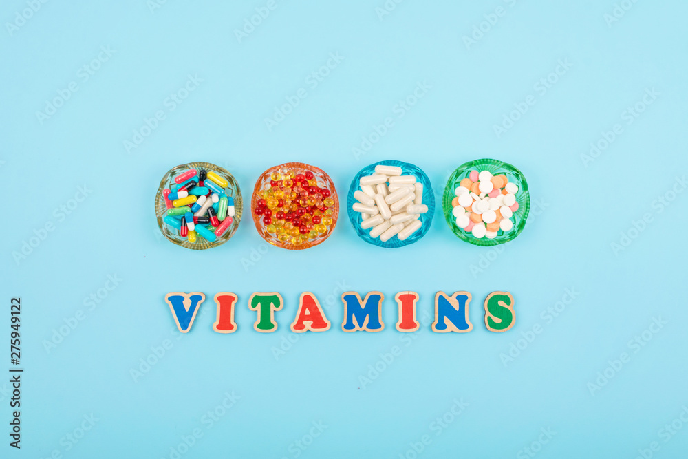 Different colorful pills and capsules in round plate and multicolored word VITAMINS on blue background. Nutrition supplement, drug, pharmaceutic concept