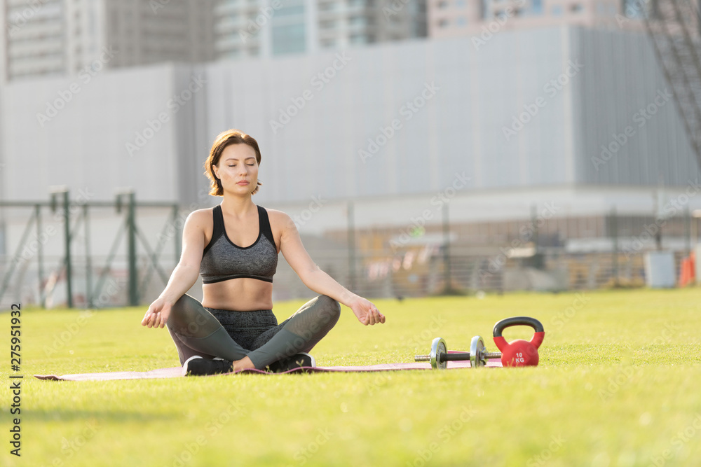 Fototapeta premium Beautiful caucasian brunette fitness model sits in a lotus cross legged position on a pink yoga mat in a open park on a bright sunny day