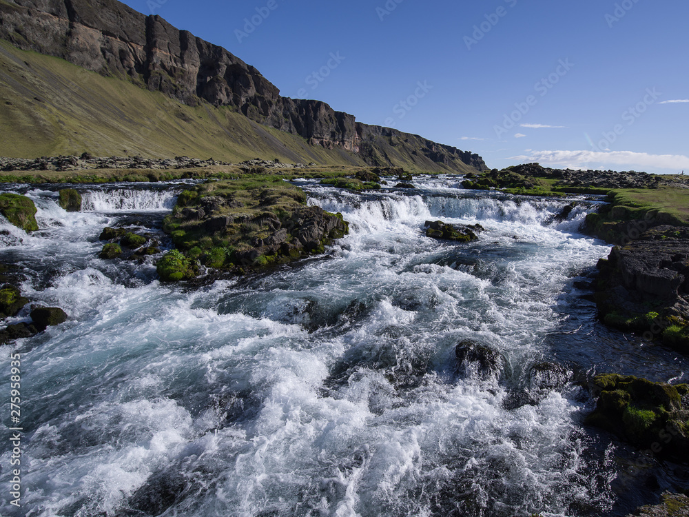 Summer in Iceland.  Rapids of fresh clear water in the center of the picture, grassy banks and volcanic rocks above. Mostly clear blue sky with some clouds on horizon