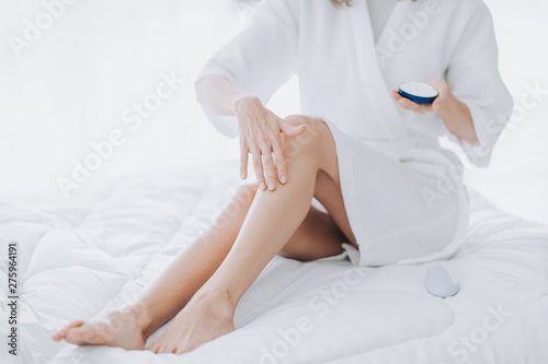 Woman apply moisturizer on her legs while seats on the bed in the morning