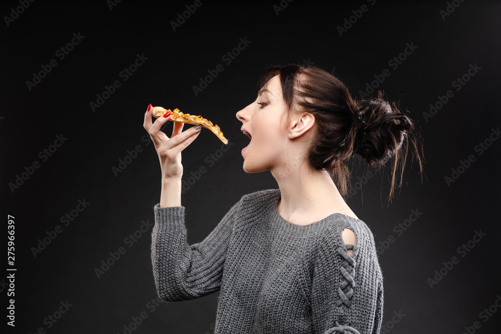Side view portrait of brunette young girl in white sweater with tail biting delicious piece of Italian pizza. Isolate on black. Fast food concept. Side view portrait of brunette young girl in white sweater with tail biting delicious piece of Italian pizza. Isolate on black. Fast food concept.