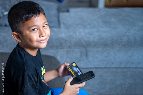 Yong Asian Children age 7-10 Years old with yellow skin, holding black credit card and black mobile phone on the sofa using his parent credit card to buy item online without permission. photo