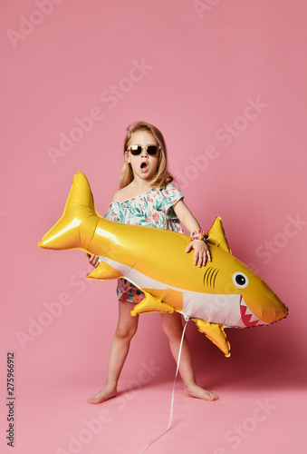 little girl, dressed in a summer dress, wears a balloon in the shape of a yellow shark-fish, celebrates a holiday, smiles broadly, stands against a pink background, is in a good mood.